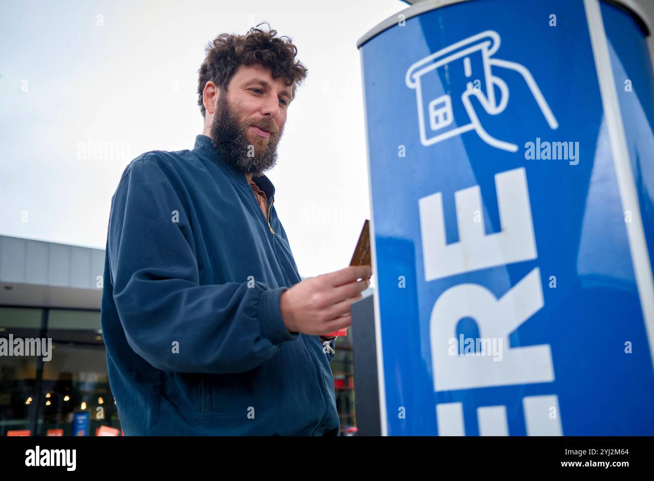 Lächelnder Mann mit Bart, der sein Smartphone benutzt, während er neben einer blauen Ladestation für Elektrofahrzeuge steht, Brüssel, Belgien Stockfoto