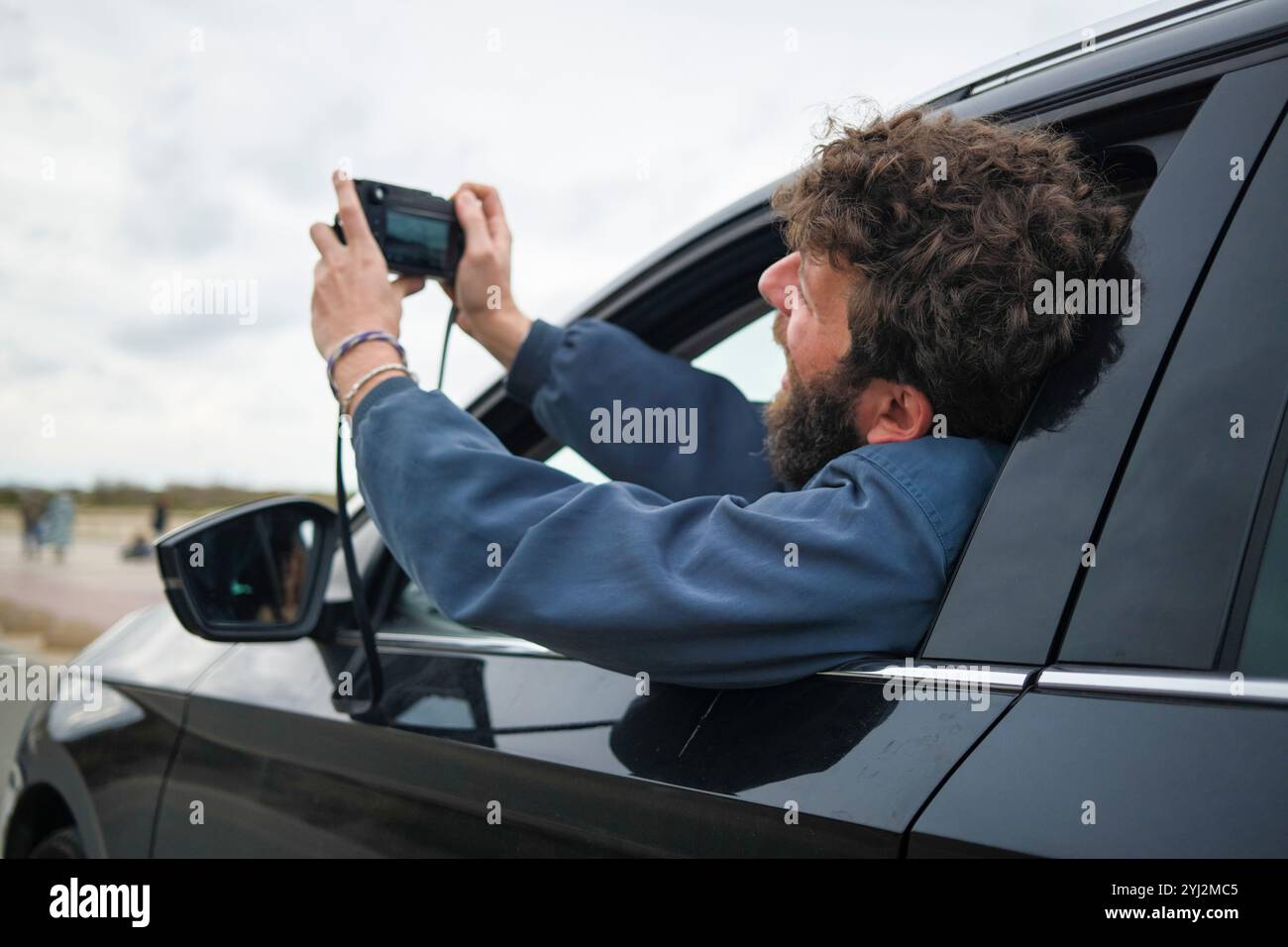 Erwachsener Mann mit lockigem Haar, der ein Foto mit seinem Smartphone aus dem Fenster eines geparkten Autos macht. Stockfoto