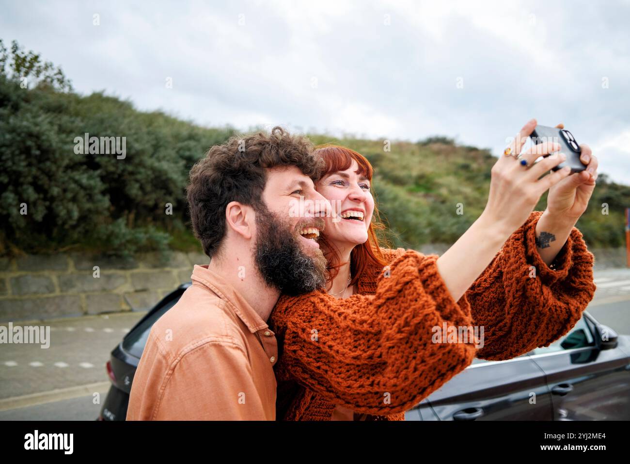 Fröhliches Paar macht ein Selfie im Freien mit einer Digitalkamera, beide lächeln breit mit einer Straße und Grün im Hintergrund. Stockfoto