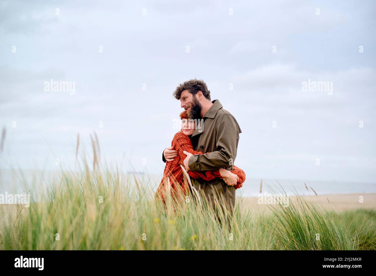 Ein Paar umarmt sich warm in hohem Gras vor einer ruhigen Strandkulisse, die Zuneigung und Ruhe vermittelt, Belgien Stockfoto