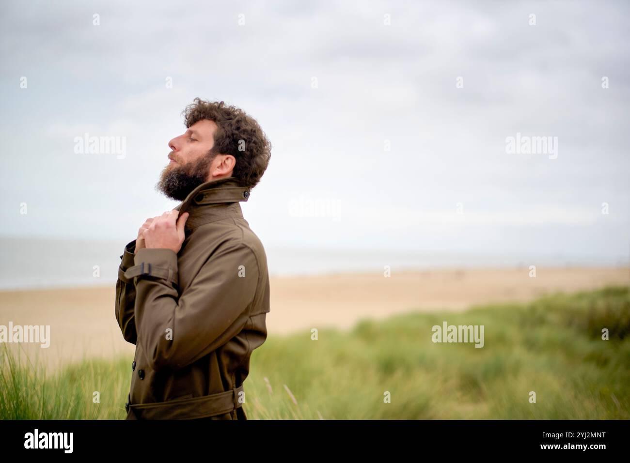 Bärtiger Mann in einer Jacke, der in grasbewachsenen Dünen steht und mit einem besinnlichen Ausdruck in den Himmel blickt, Belgien Stockfoto