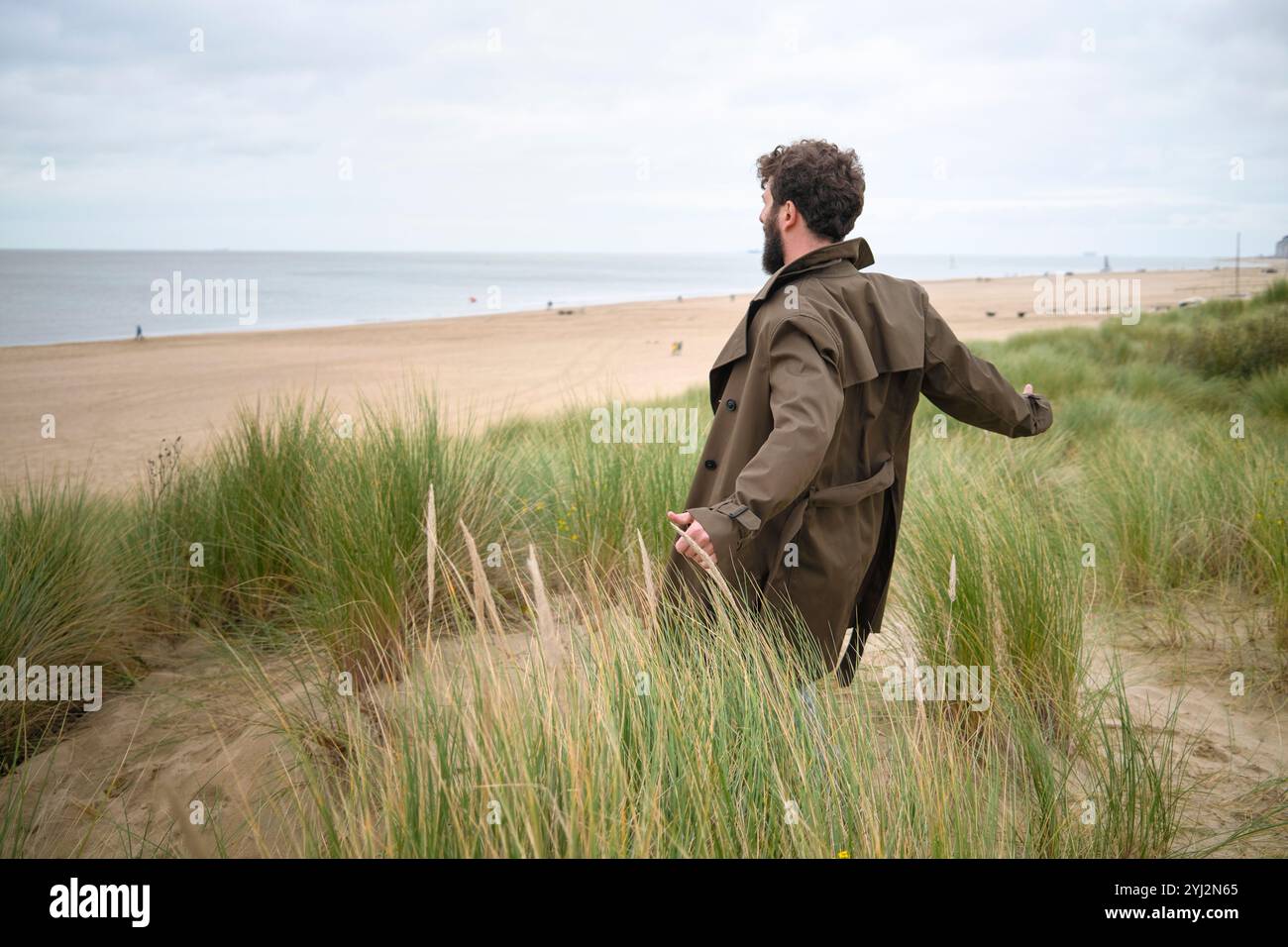 Mann in beigefarbenem Mantel, der durch Küstendünen mit grünem Gras und bewölktem Himmel geht, Belgien Stockfoto