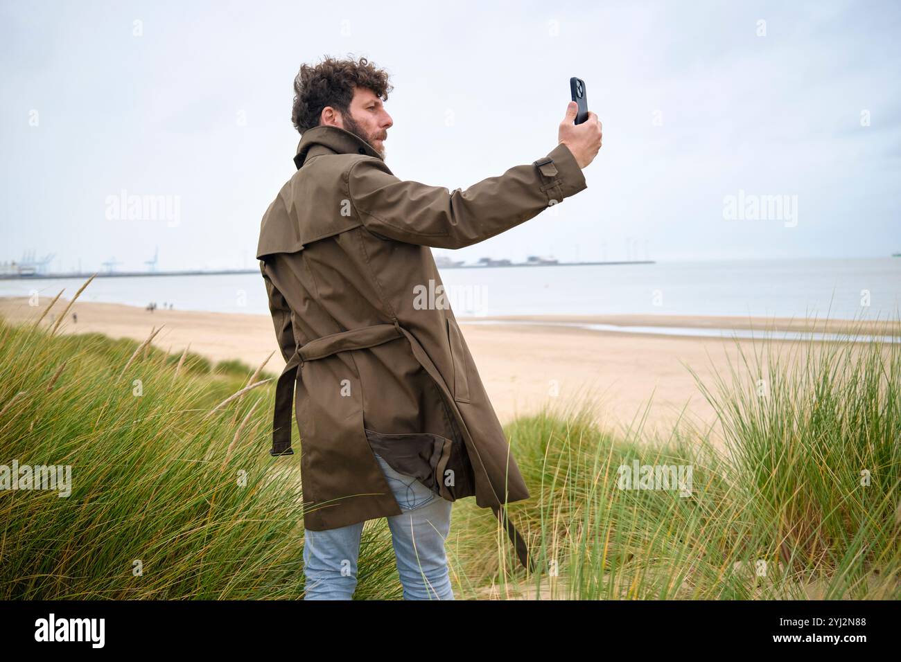 Mann in braunem Mantel, der ein Selfie mit seinem Handy macht, an einem Strand mit grasbewachsenen Dünen im Vordergrund und bedecktem Himmel, Belgien Stockfoto