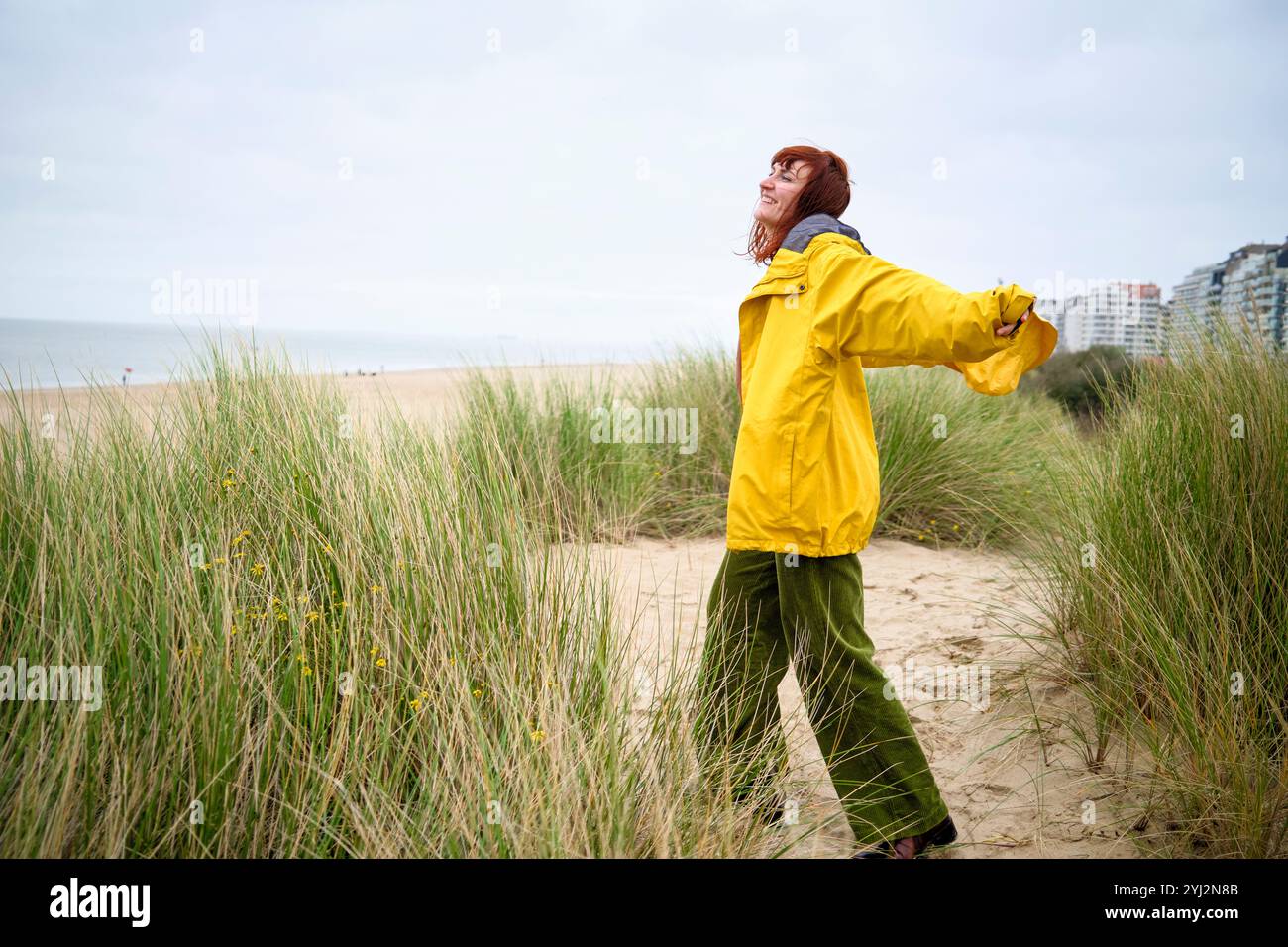 Frau in gelbem Regenmantel genießt eine Brise auf einer grasbewachsenen Sanddüne mit Gebäuden in der Ferne, Belgien Stockfoto