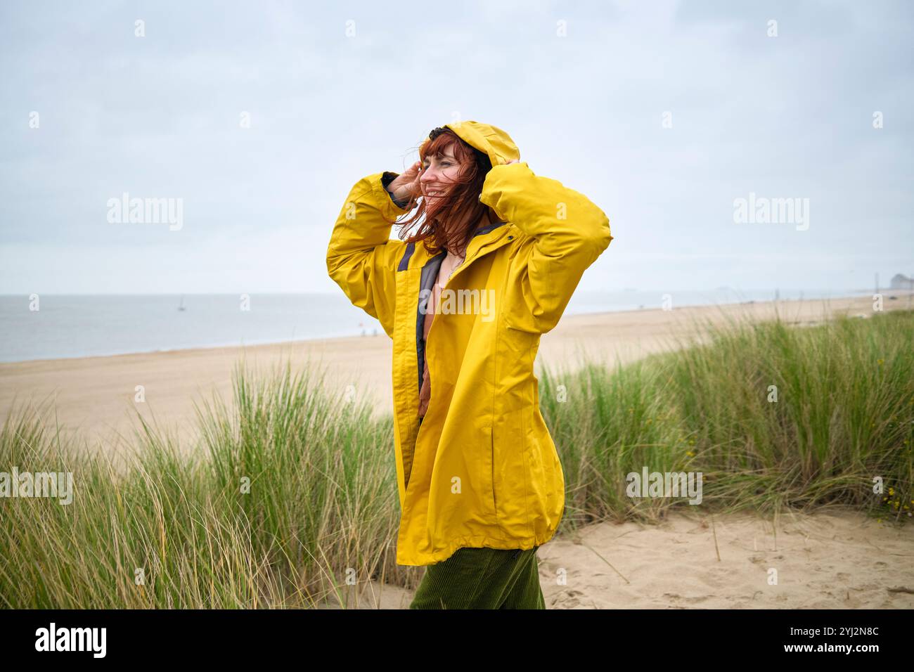 Eine fröhliche Frau in gelbem Regenmantel steht am Strand mit grasbewachsenen Dünen, lächelt und hält ihre Haare an einem luftigen Tag in Belgien Stockfoto