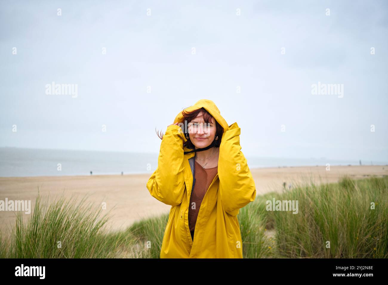 Frau in gelbem Regenmantel steht an einem luftigen Strand mit Gras im Vordergrund, Belgien Stockfoto