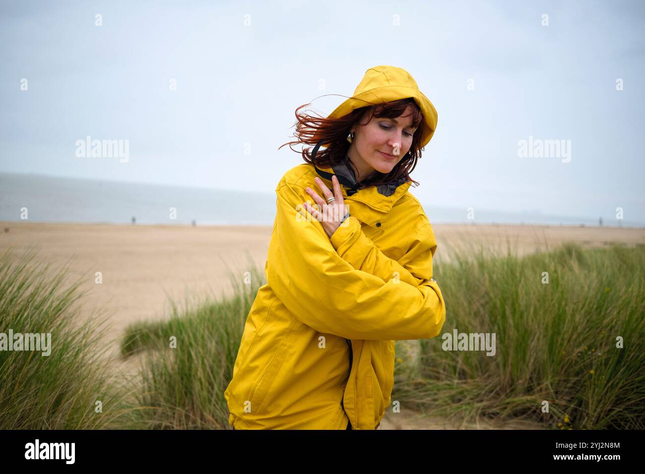 Frau in gelbem Regenmantel steht an einem Strand mit grasbewachsenen Dünen und schließt ihre Augen vor dem Wind, Belgien Stockfoto