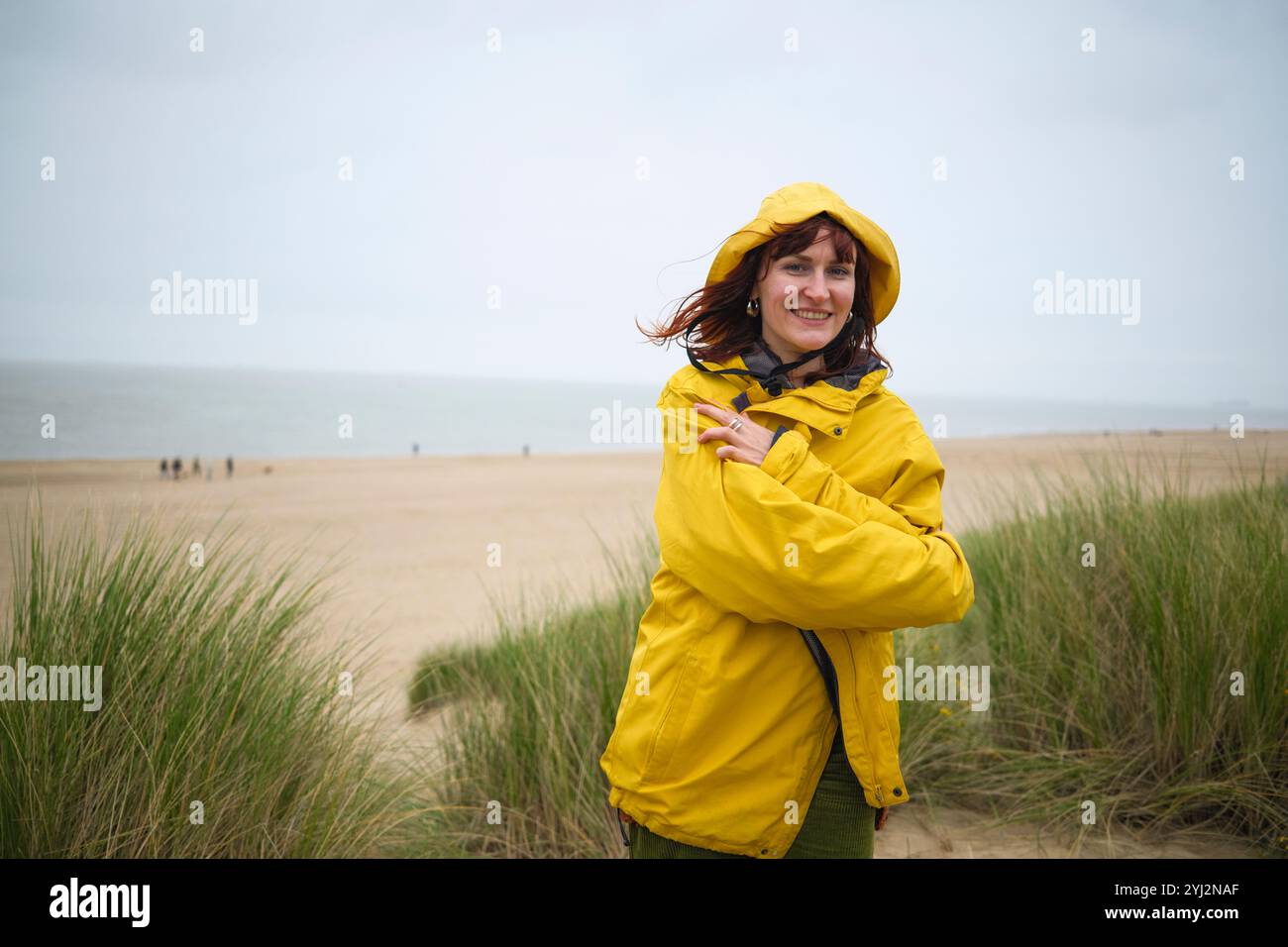 Lächelnde Frau in gelbem Regenmantel steht an einem Strand mit Dünen und bedecktem Himmel, Belgien Stockfoto