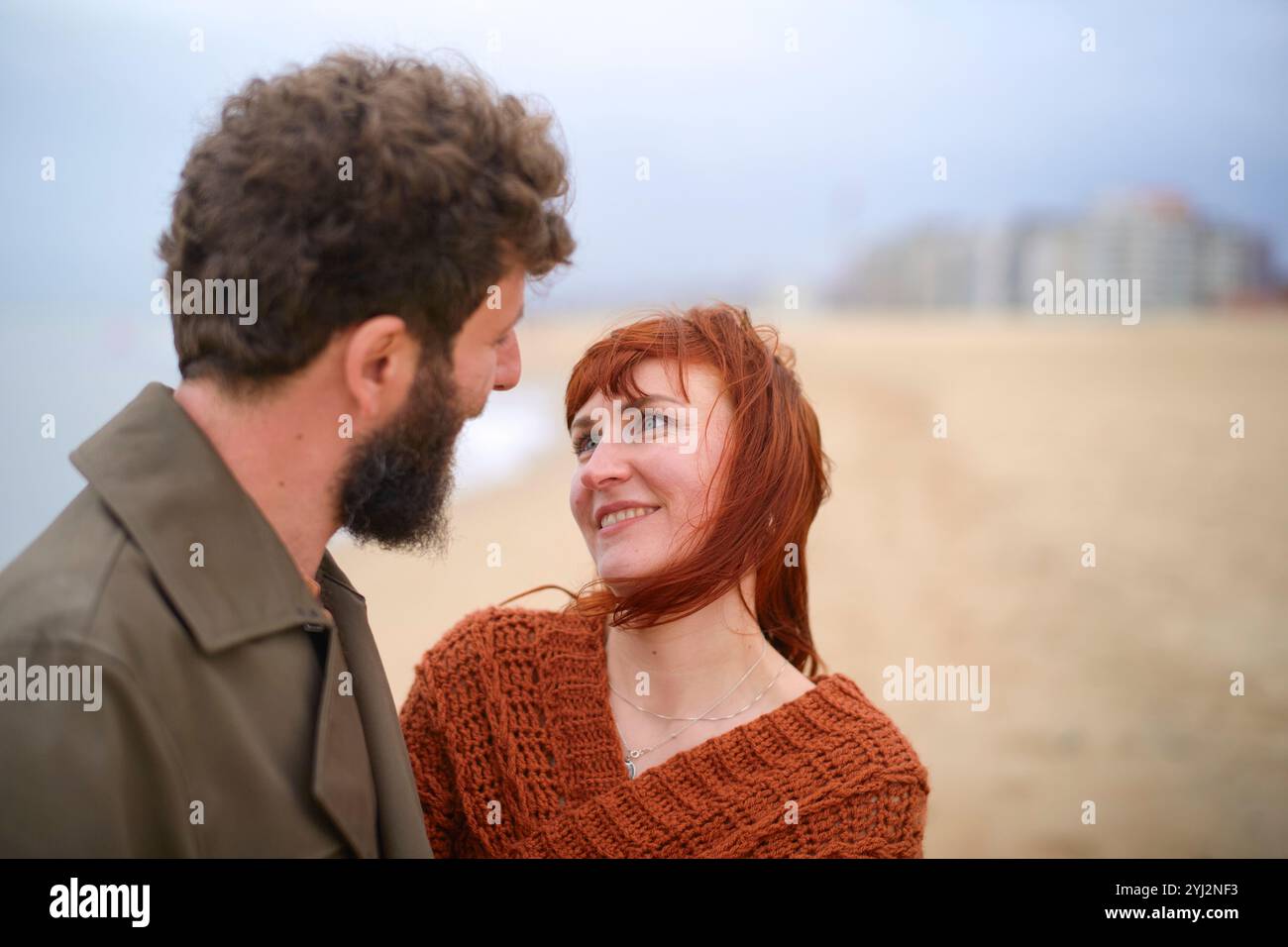 Eine Frau mit roten Haaren schaut liebevoll einen bärtigen Mann vor einer verschwommenen Strandkulisse an, Belgien Stockfoto