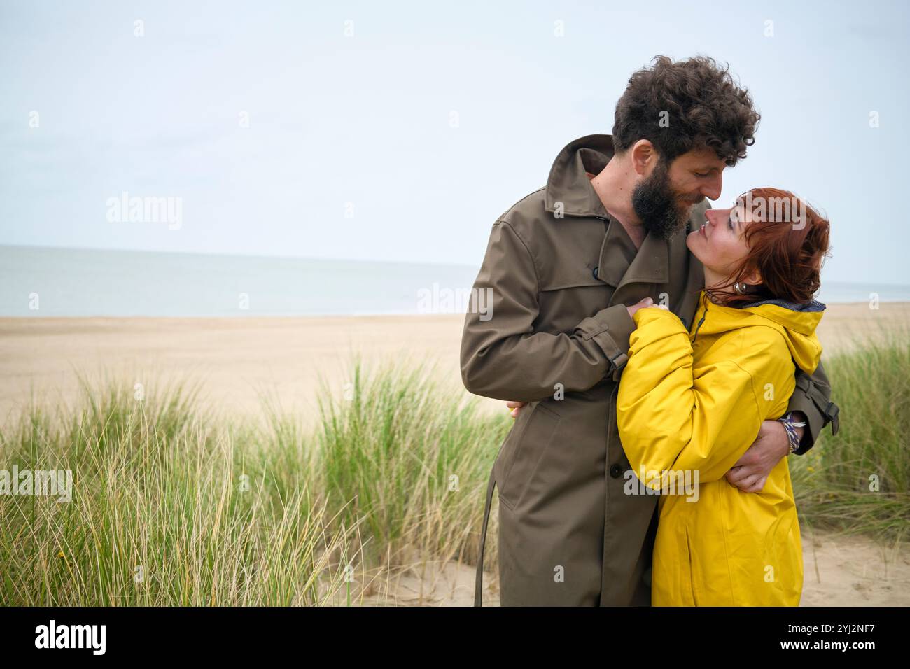 Ein romantisches Paar, das sich an einem Strand mit bewölktem Himmel umgibt, der Mann im Trenchcoat und die Frau im gelben Regenmantel, Belgien Stockfoto