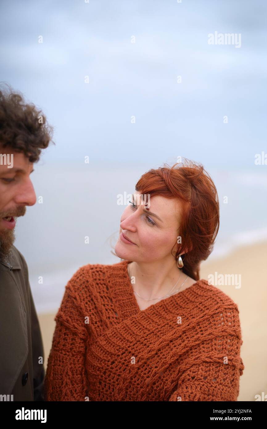 Frau in einem rostfarbenen Pullover, der einen Mann vor einer ruhigen Strandkulisse ansieht, Belgien Stockfoto