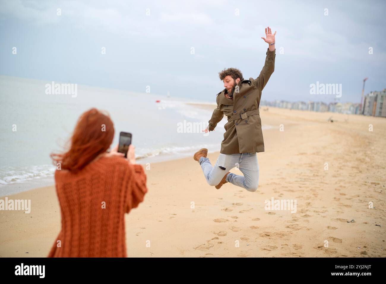 Mann im Luftsprung an einem Sandstrand, der von einer Frau mit Smartphone fotografiert wird, Belgien Stockfoto