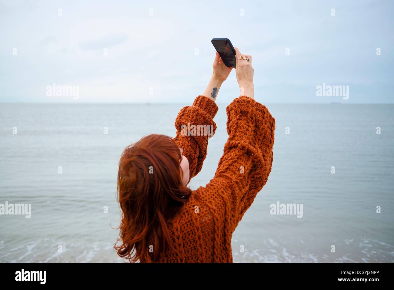 Frau in einem orangefarbenen Pullover, die mit ihrem Smartphone ein Foto vom Meereshorizont macht, Belgien Stockfoto