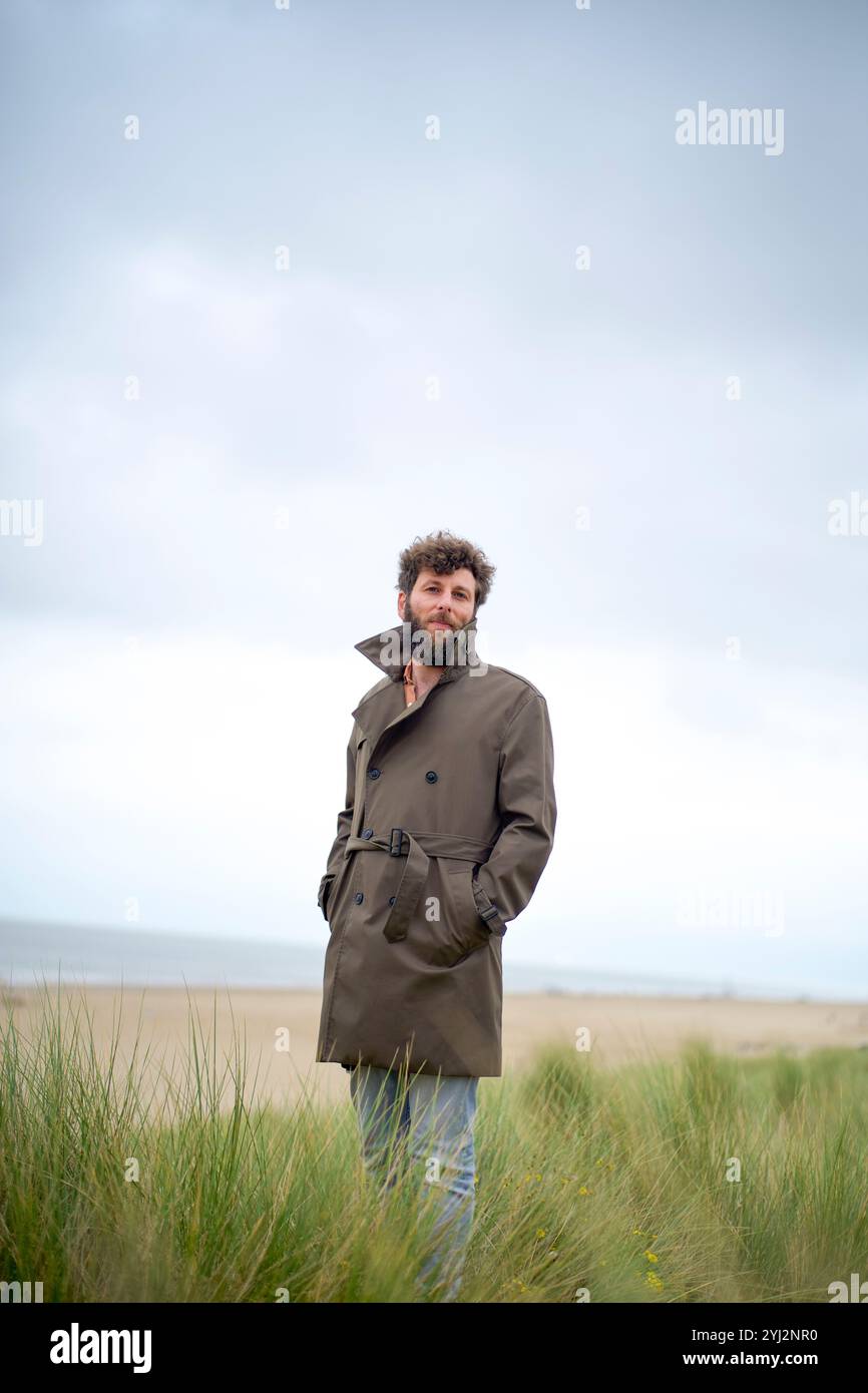 Mann in braunem Mantel auf einem Feld mit hohem Gras und bewölktem Himmel im Hintergrund, Belgien Stockfoto