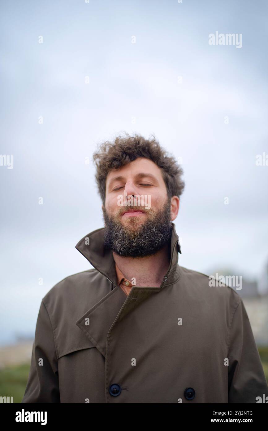 Mann mit lockigen Haaren und Bart, der die frische Luft mit geschlossenen Augen und zufriedenem Ausdruck genießt, in braunem Mantel, Belgien Stockfoto