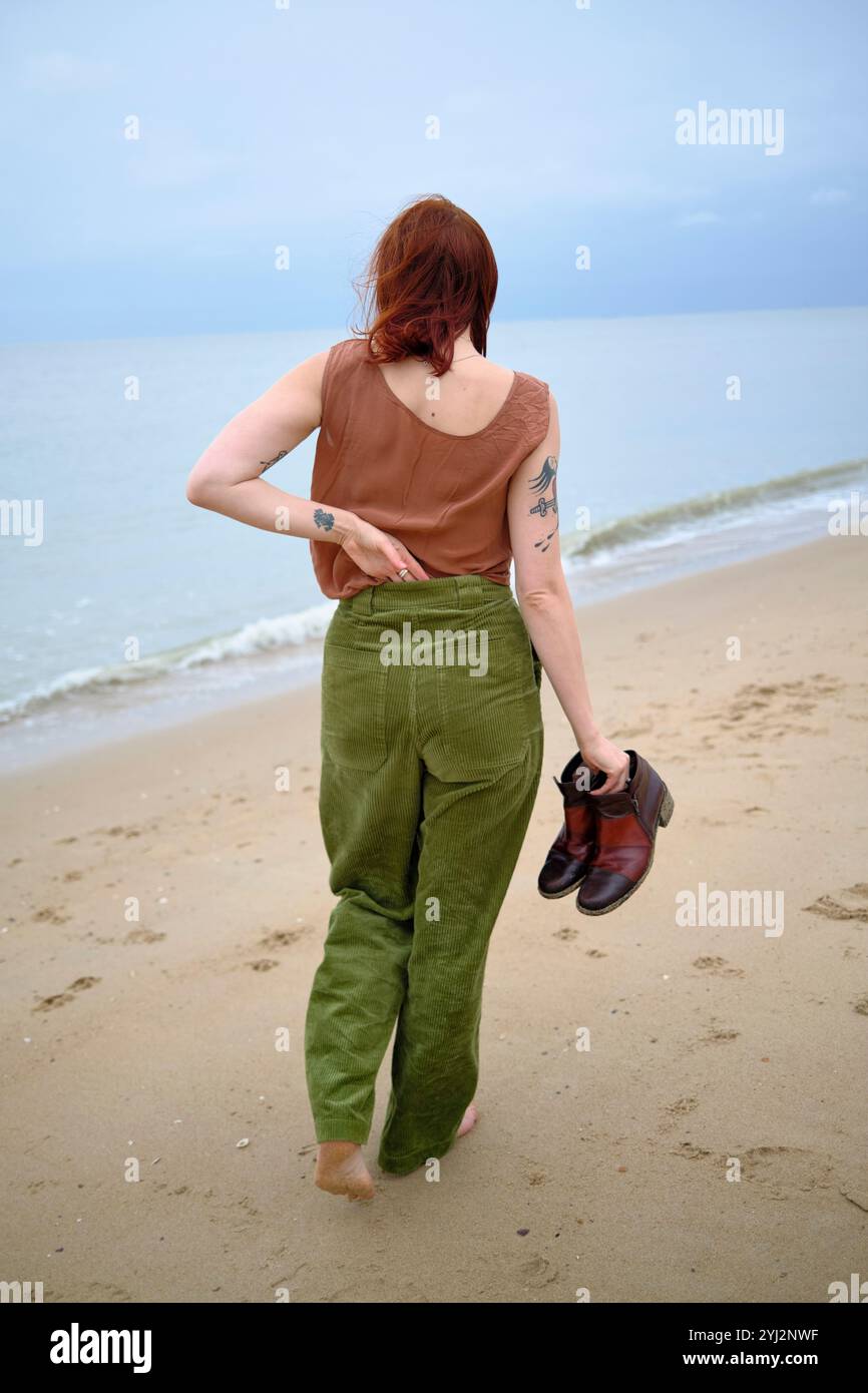 Frau mit Tätowierungen läuft barfuß an einem Sandstrand und hält ihre Schuhe in der Hand, Belgien Stockfoto