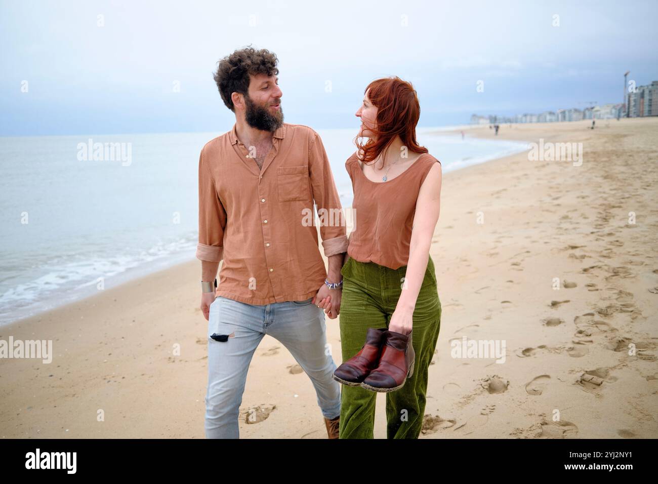 Ein bärtiger Mann in einem sandfarbenen Hemd und eine Frau mit rotem Haar, die ein Tanktop trägt, lächeln einander an, während sie barfuß an einem Strand in Belgien spazieren Stockfoto