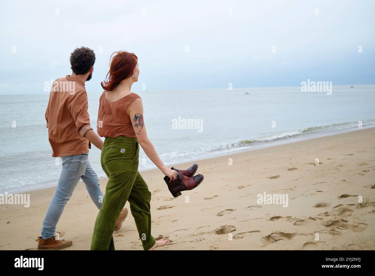 Paare halten Hände, während sie barfuß an einem Sandstrand mit Schuhen in der Hand laufen, mit einem ruhigen Meer und bewölktem Himmel im Hintergrund, Belgien Stockfoto Paare halten Hände, während sie barfuß an einem Sandstrand mit Schuhen in der Hand laufen, mit einem ruhigen Meer und bewölktem Himmel im Hintergrund, Belgien Stockfoto