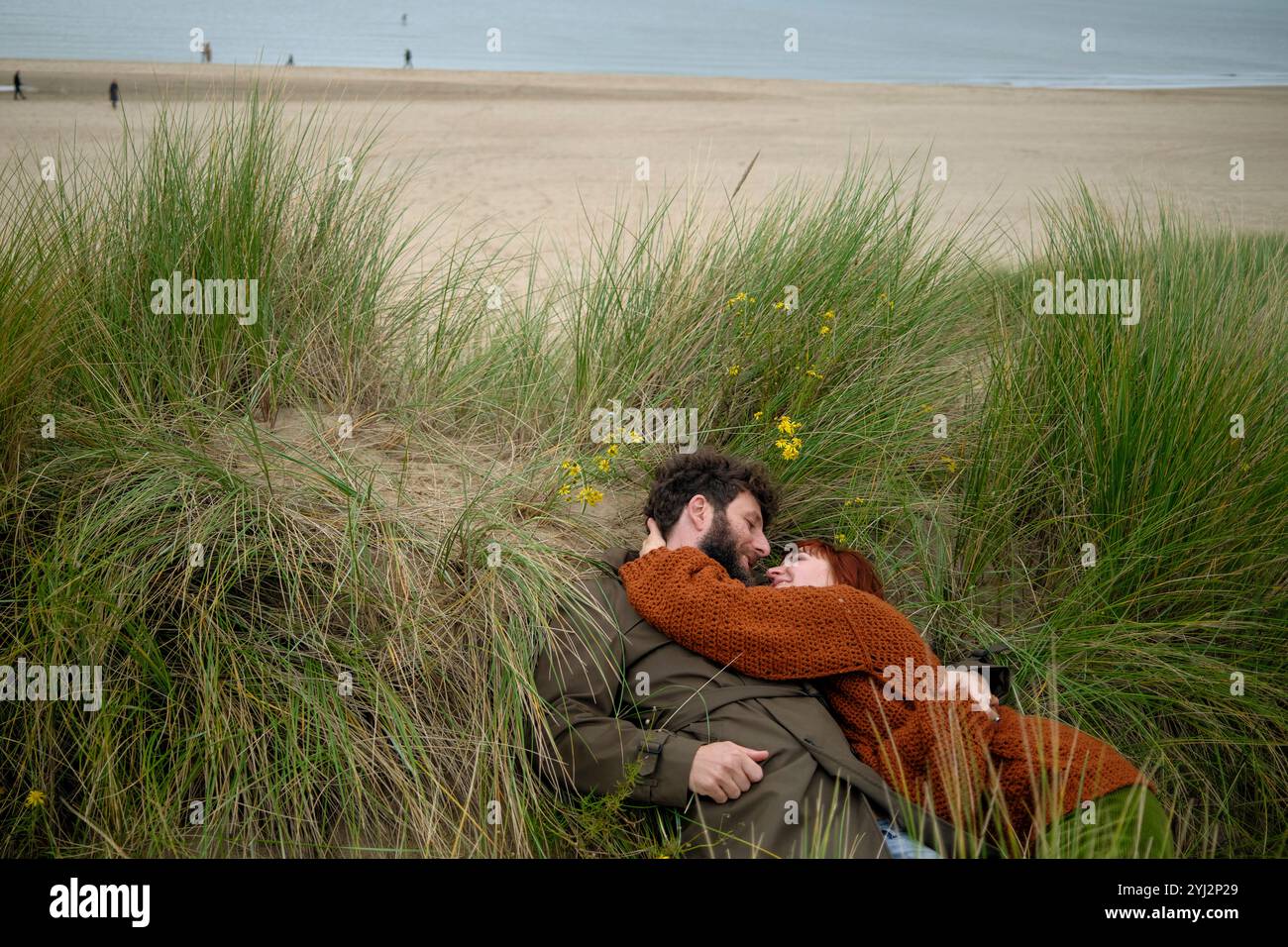 Ein Paar umarmt sich im Strandgras mit gelben Blumen, einem Sandstrand und weit entfernten Figuren im Hintergrund, Belgien Stockfoto