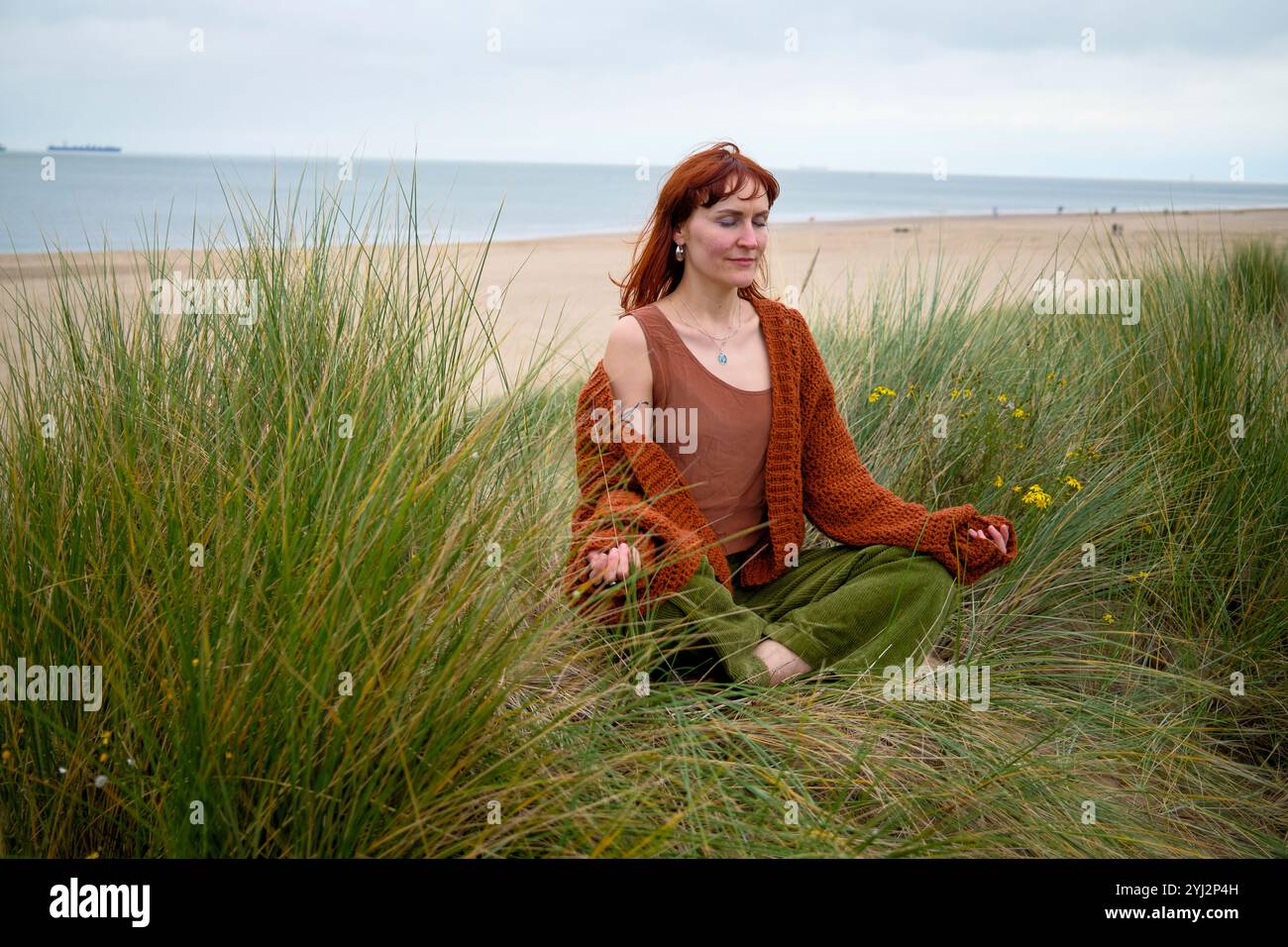 Frau, die in hohem Gras mit einer ruhigen Strandkulisse meditiert, Belgien Stockfoto