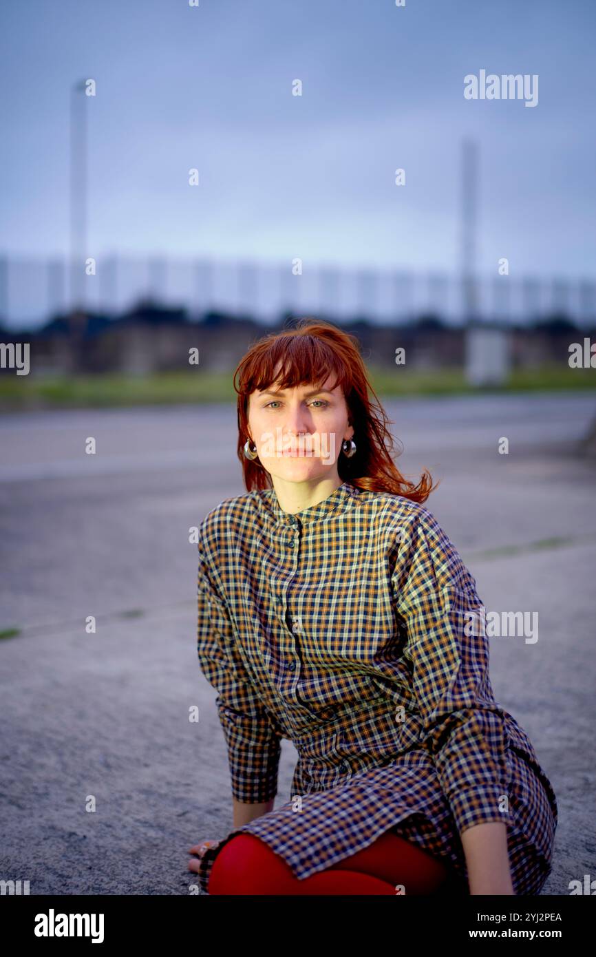 Frau mit rotem Haar, die ein kariertes Hemd und eine rote Hose trägt, sitzt in der Abenddämmerung auf einem Betonboden Stockfoto
