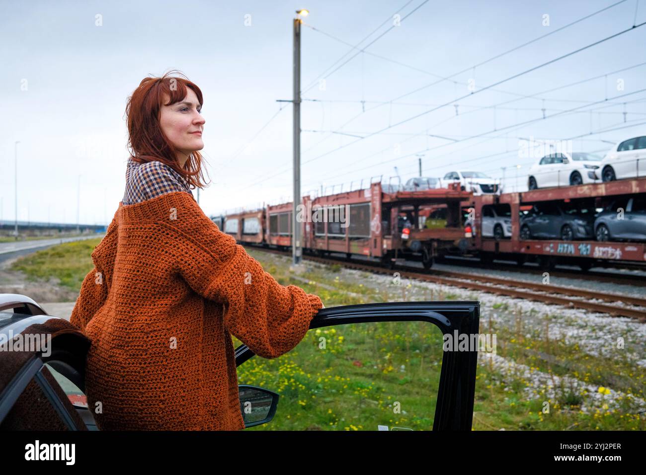 Frau in einem orangefarbenen Pullover, lehnt sich an eine Autotür, lächelnd, mit Eisenbahnwaggons und bedecktem Himmel im Hintergrund, Belgien Stockfoto