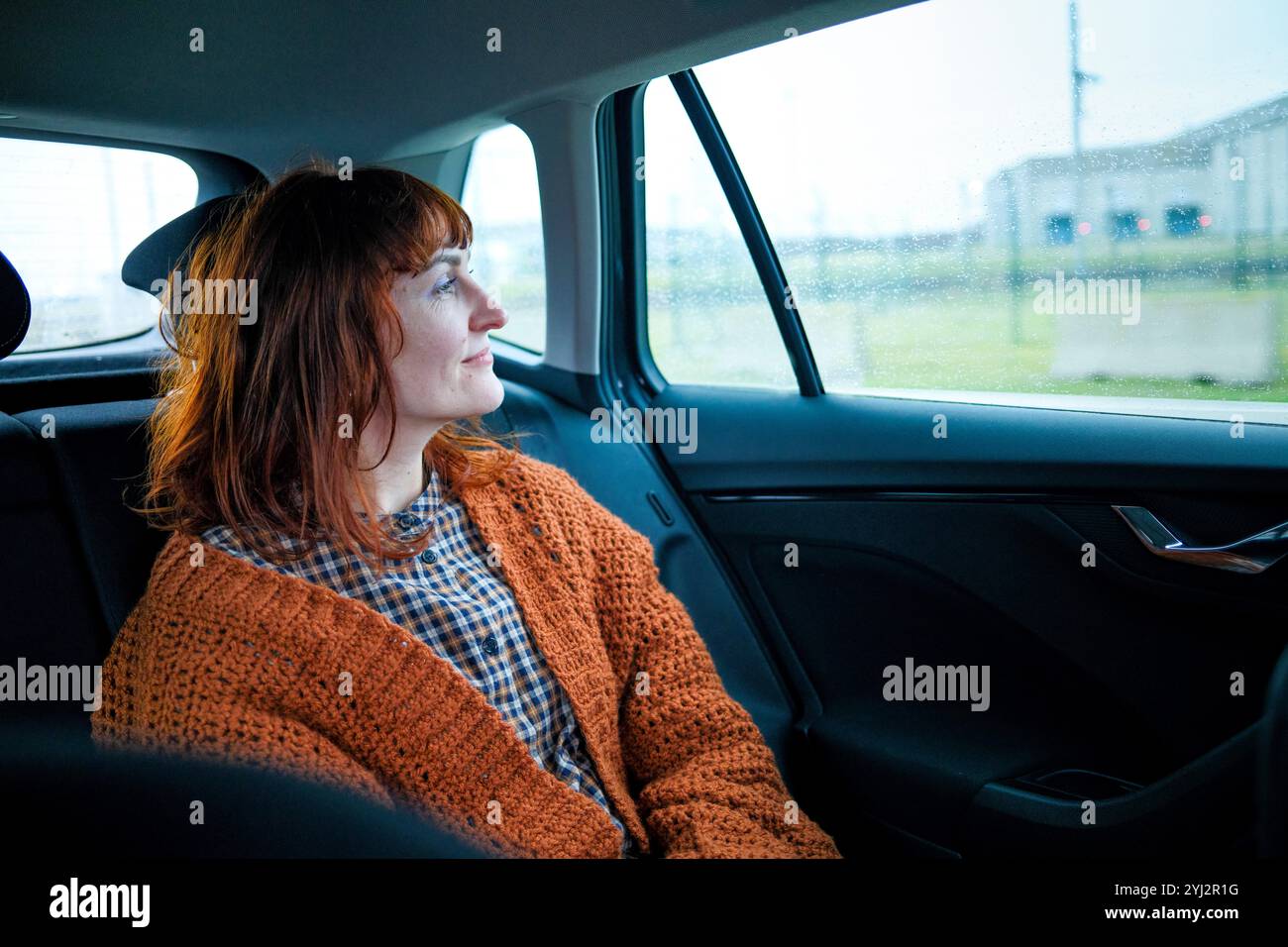 Frau in orangefarbenem Pullover, die an einem regnerischen Tag eine Autofahrt genießt und mit einem dezenten Lächeln aus dem Fenster blickt. Stockfoto