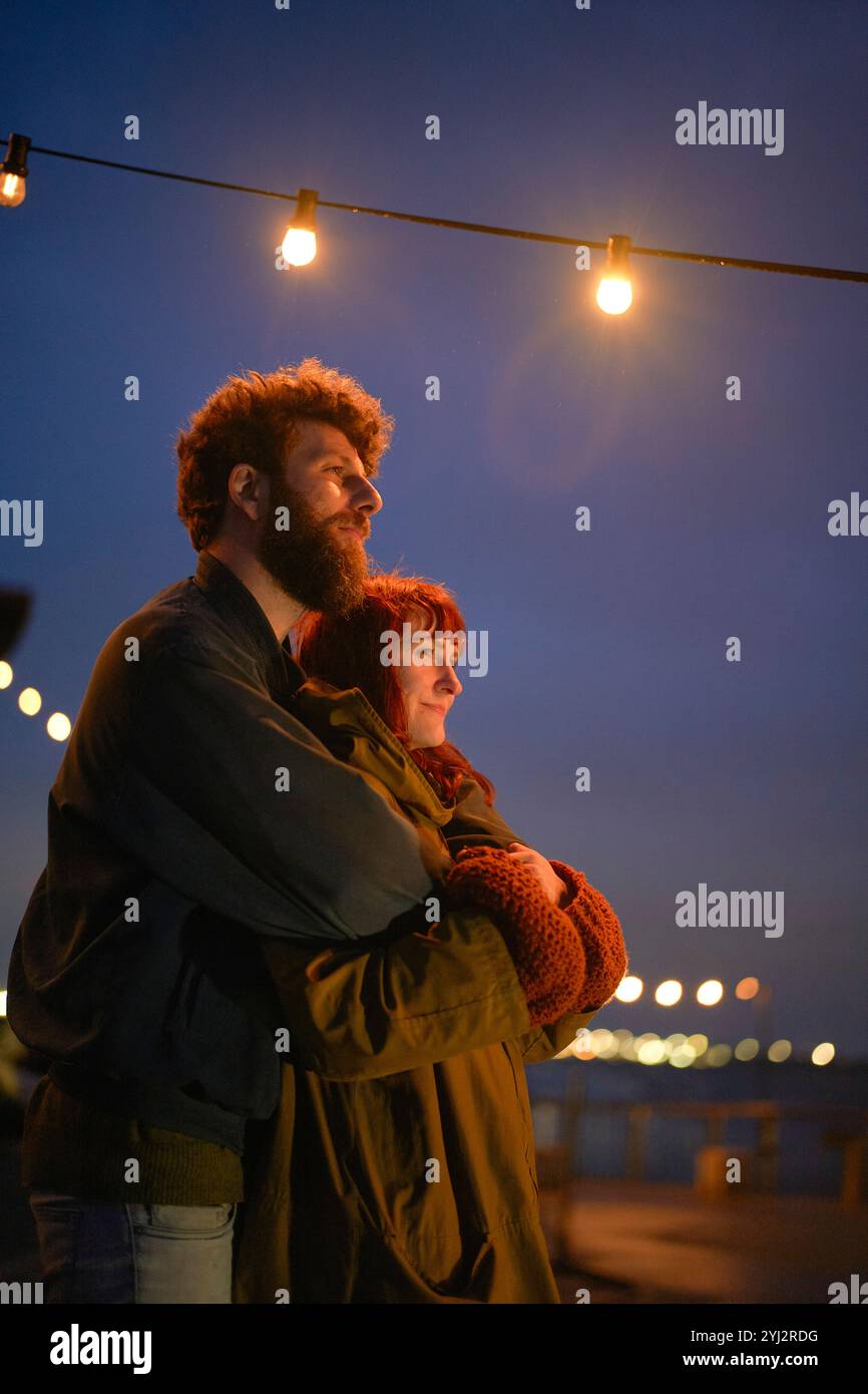 Paare, die sich unter Abendlichtern an einem Pier am Wasser mit einem ruhigen Gesichtsausdruck umarmen, Belgien Stockfoto