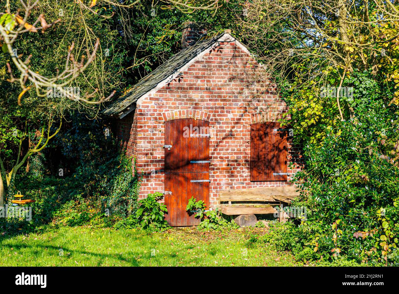 Kleiner Lagerschuppen im Garten des Bauernhauses, Backsteinmauern, Tür und Fenster mit Holzschirm geschlossen, viele grüne Pflanzen und Bäume, sonniger Tag Stockfoto