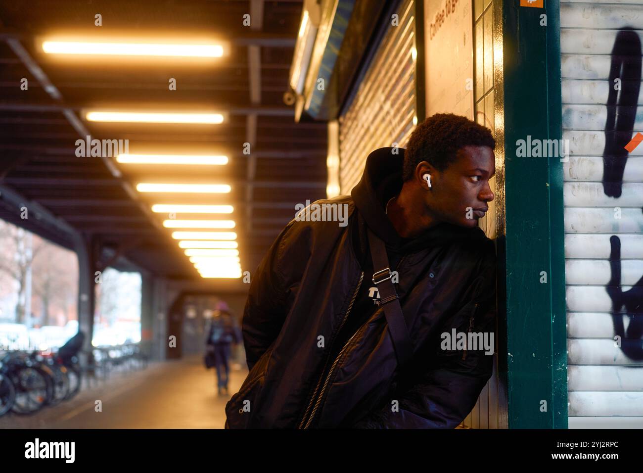 Ein junger Mann mit Ohrhörern lehnt sich an eine gekachelte Wand unter dem Schein von Deckenleuchten in einer U-Bahn-Station in Berlin Stockfoto