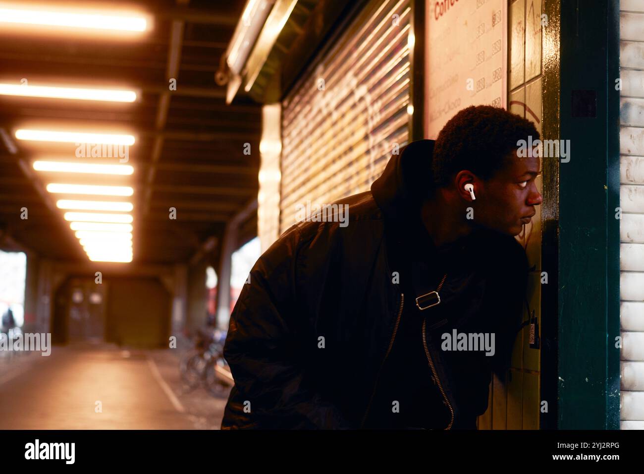 Ein junger Mann mit Ohrhörern lehnt sich an eine gekachelte Wand unter dem Schein von Deckenleuchten in einer U-Bahn-Station in Berlin Stockfoto