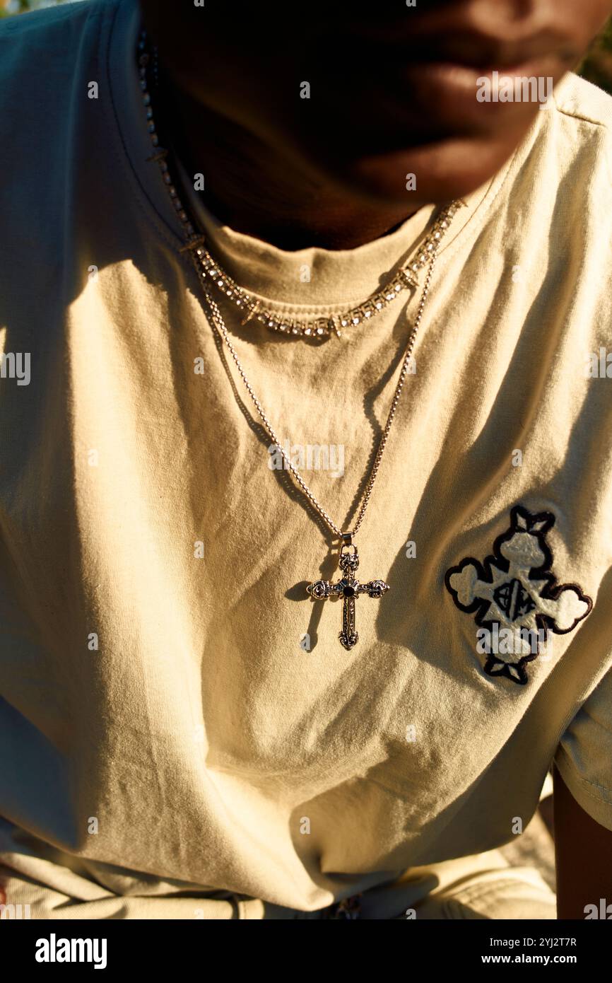 Sonnenlicht strahlt auf eine Person, die ein beiges T-Shirt und mehrlagige Halsketten trägt, von denen eine einen Kreuzanhänger trägt. Stockfoto