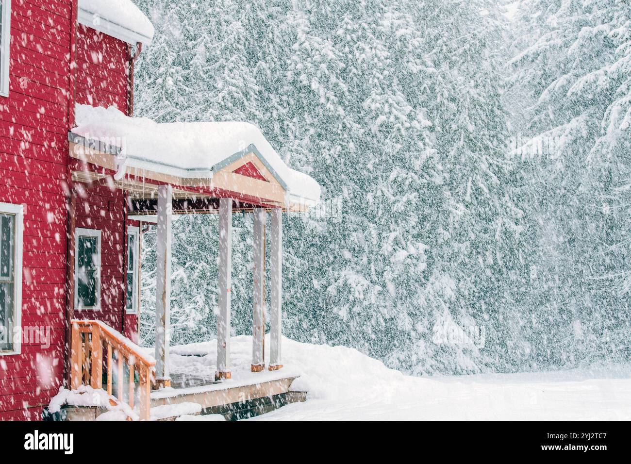 Rote Hütte mit einer Veranda, umgeben von einem schweren Schneesturm vor einem Hintergrund von schneebedeckten Bäumen. Stockfoto