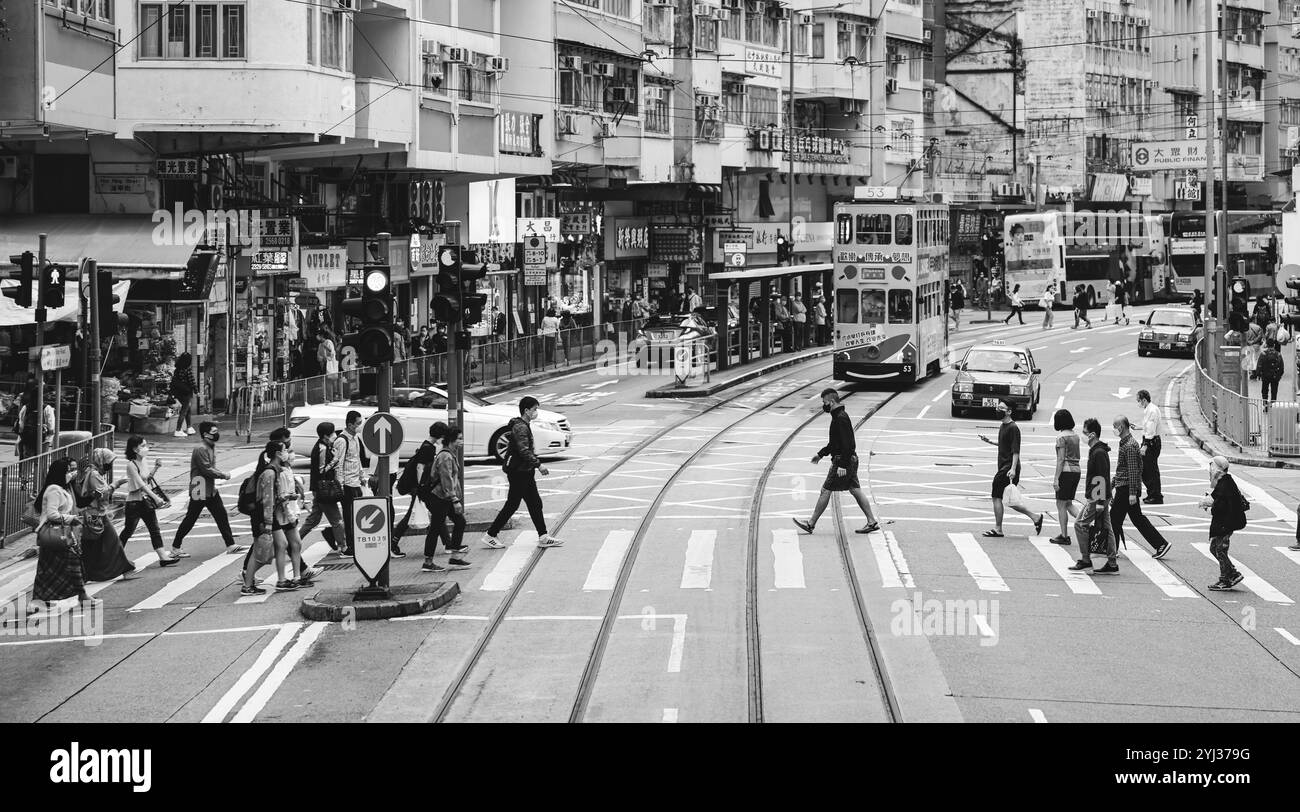 Fußgänger fahren an einem typischen Tag in Hongkong durch eine belebte Straße voller Straßenbahnen, Geschäfte und Ampeln. Stockfoto