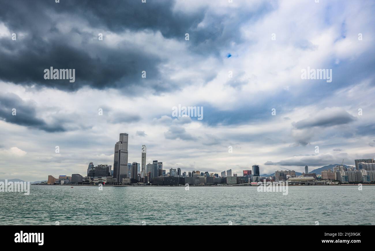 Ein weitläufiger ausblick zeigt die moderne Skyline von Hongkong entlang der Uferpromenade, die sich im Wasser spiegelt, während sich Wolken über dem Wasser sammeln. Stockfoto