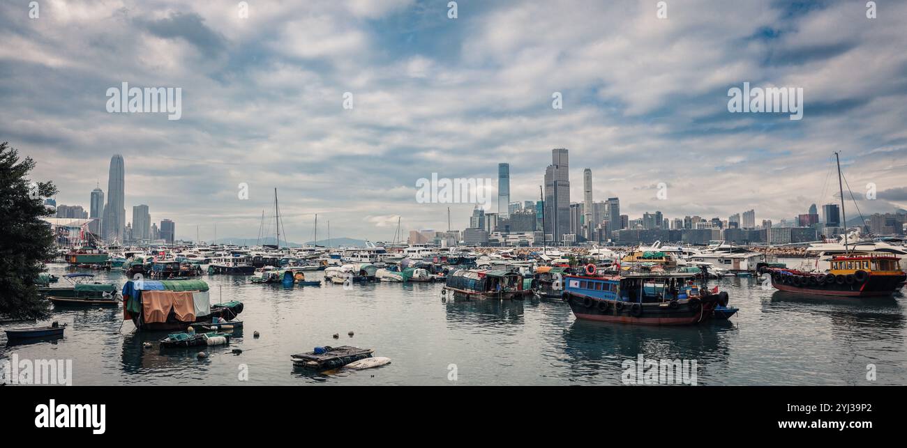 Ein weitläufiger blick auf Hongkong zeigt die Skyline der Stadt entlang eines belebten Hafens voller traditioneller Boote und dramatischer Wolkenformationen. Stockfoto