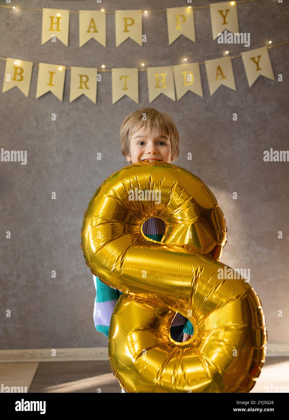 Glücklicher 8-jähriger Junge, der seinen Geburtstag feiert. Der Geburtstagsjunge steht im Zimmer und hält einen goldenen Festball in Form von Nummer acht in seinen Zähnen. Geburtsdatum Stockfoto