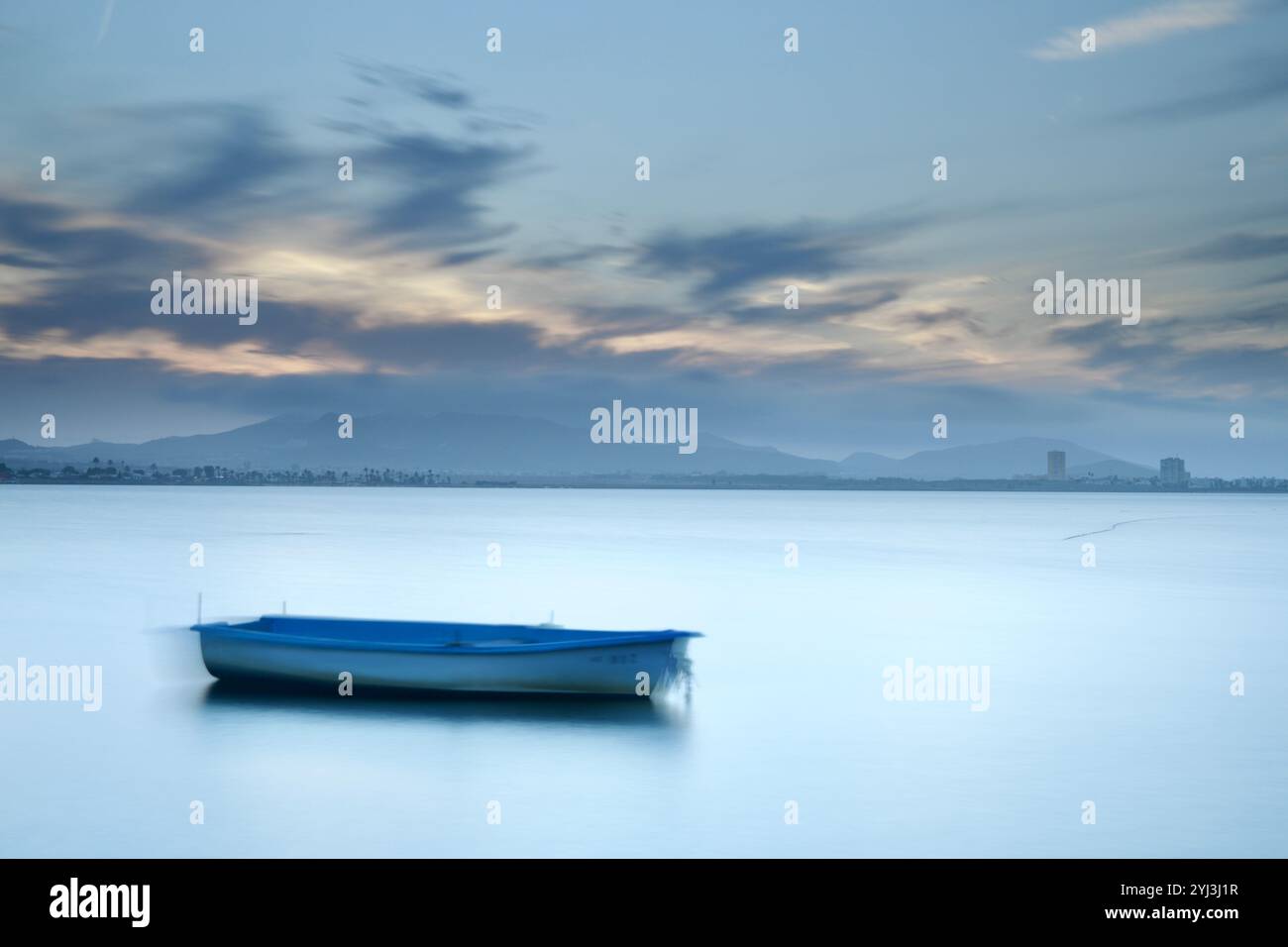Langzeitbelichtung eines kleinen Bootes, das bei Sonnenuntergang auf dem ruhigen Wasser des Mar Menor schwimmt, Region Murcia, Spanien, mit weichen Wolken und fernen Hügeln Stockfoto