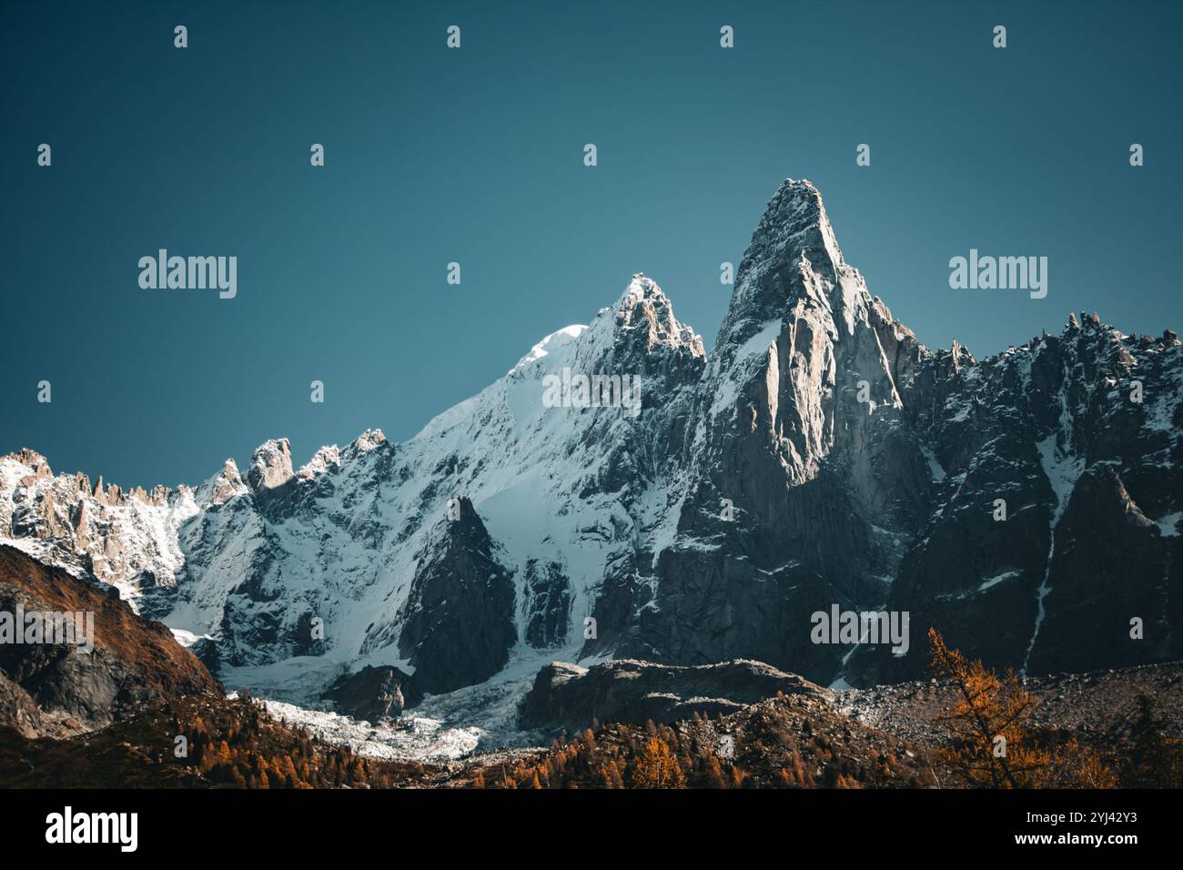 Schneebedeckte Berggipfel ragen über felsiges Gelände, beleuchtet von warmem Sonnenlicht, mit Herbstbäumen, die einen Kontrast unter einem klaren blauen Himmel bilden. Stockfoto
