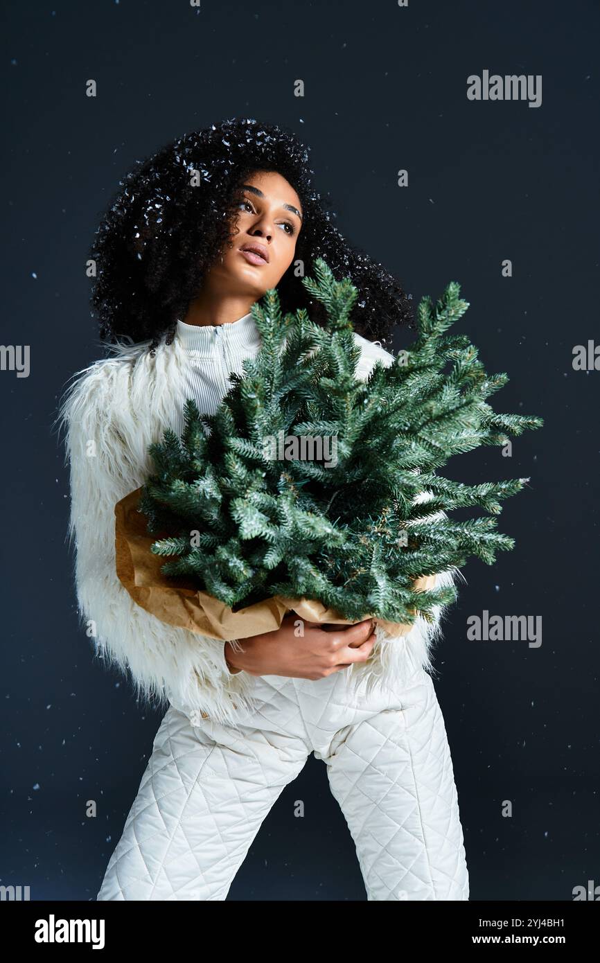 Eine modische afroamerikanische Frau in einem eleganten Skianzug und einer Kunstpelzjacke hält einen winterlich immergrünen Baum. Stockfoto