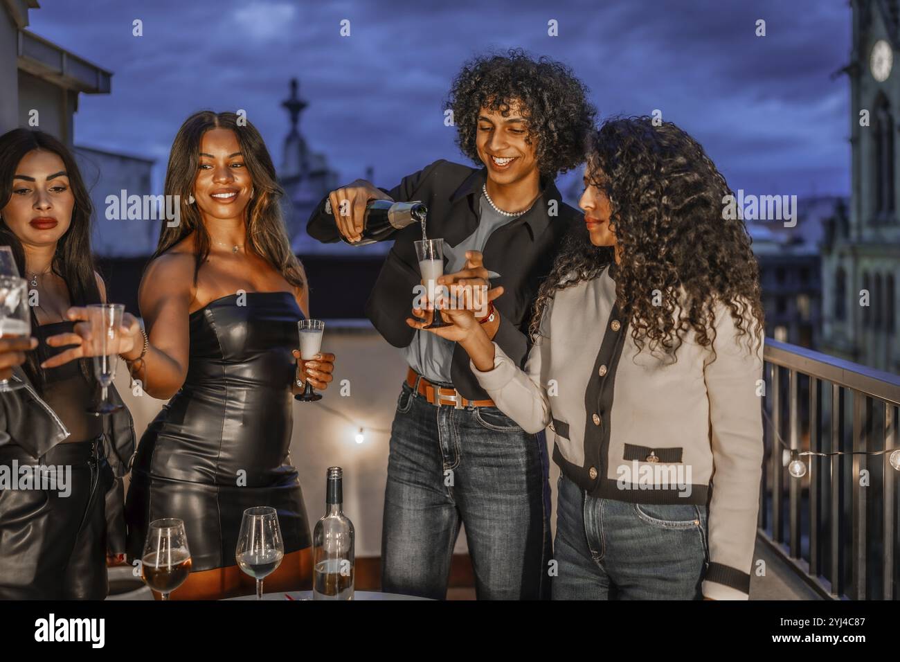 Mann, der multiethnischen Freunden Champagner in Gläsern serviert, auf einer Nachtparty auf einem Dach Stockfoto