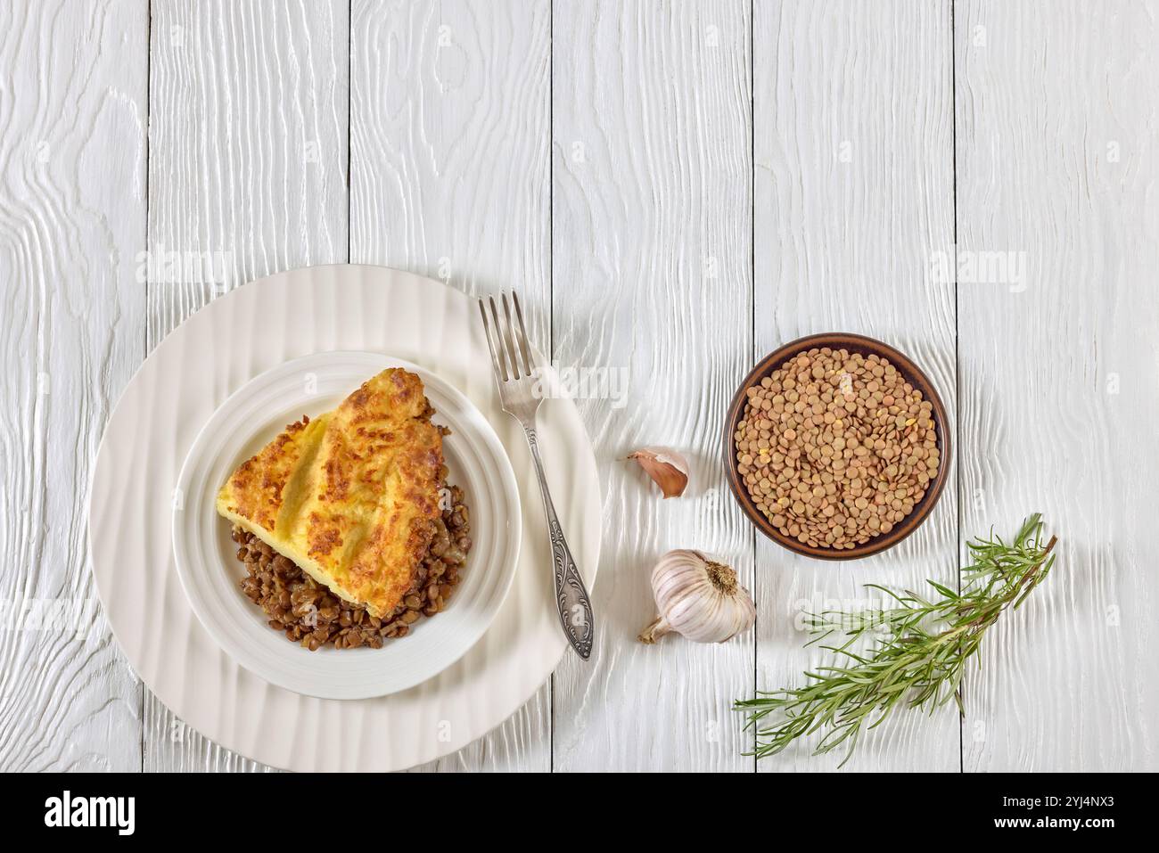 Veganer Hirtenkuchen mit gekochten Linsen, belegt mit Kartoffelpüree auf weißem Teller mit Gabel auf Holztisch mit Rosmarin, horizontale Ansicht von oben, Stockfoto