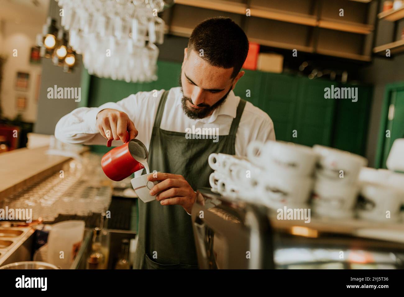 In einem warmen und einladenden Café gießt ein engagierter Barista fachmännisch Milch in eine Kaffeetasse und sorgt so für ein angenehmes Erlebnis für Gäste. Stockfoto