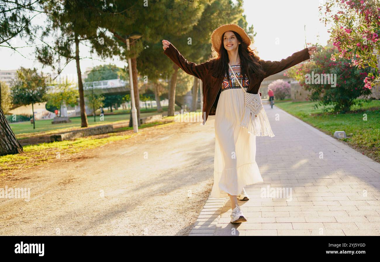 Eine freudige Frau geht in der Natur, trägt ein fließendes Outfit und trägt einen Sonnenhut Stockfoto