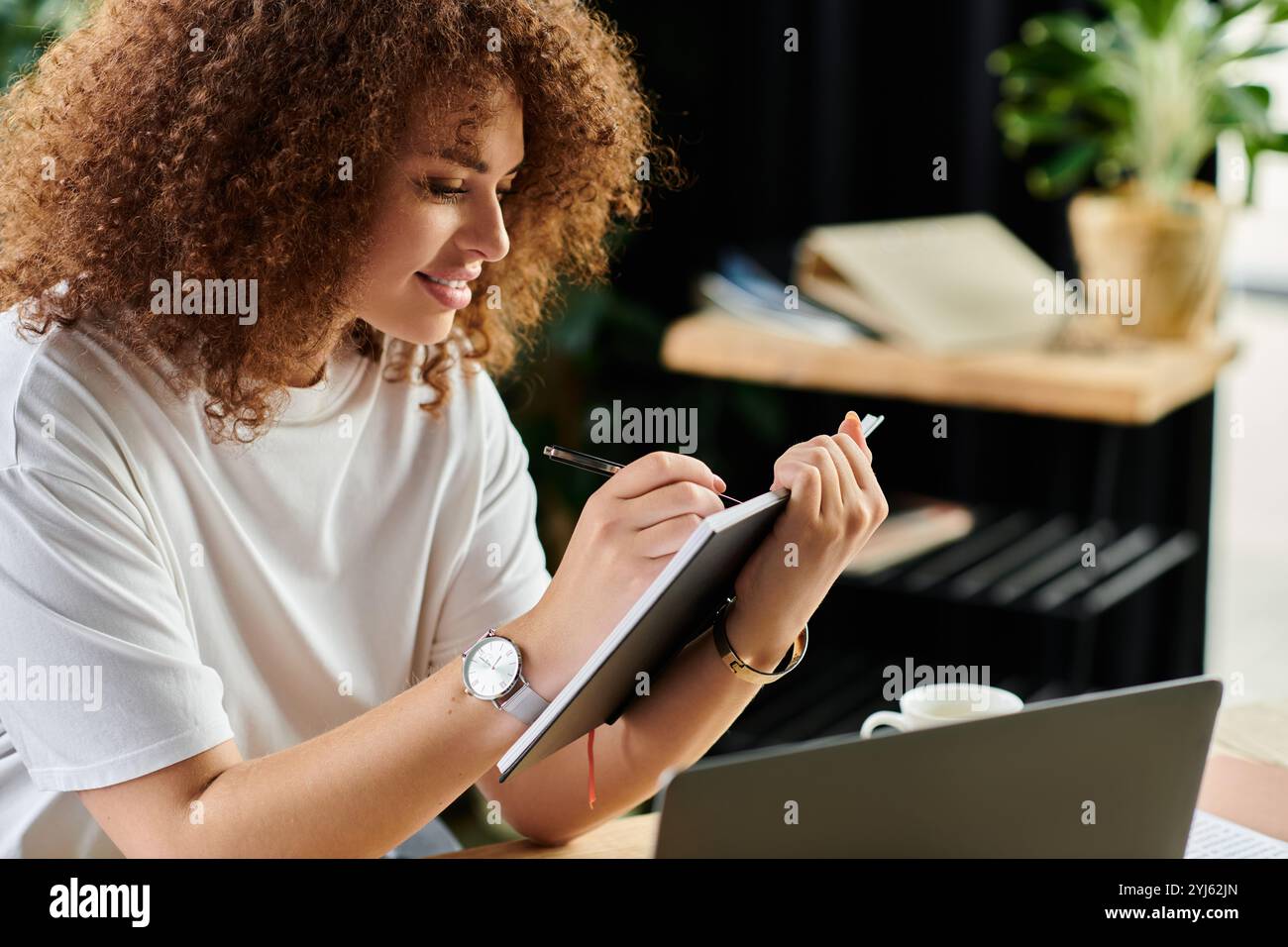 Eine Frau mit lockigen Haaren konzentriert sich auf ihr Notizbuch, die sich intensiv mit ihrer Arbeit an einem gemütlichen Schreibtisch beschäftigt. Stockfoto