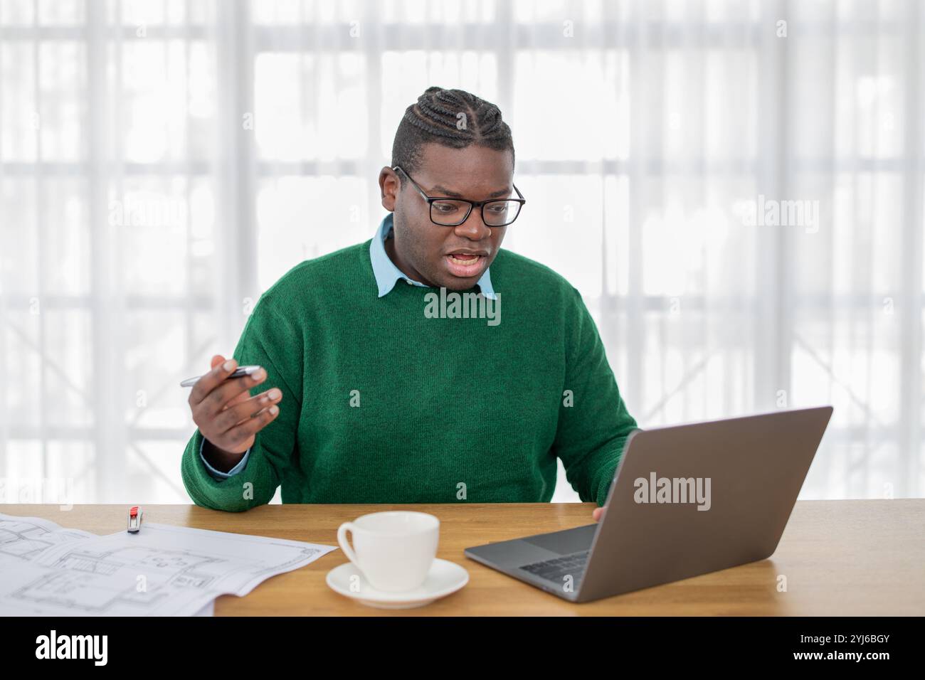 Ein schwarzer Mann in einem grünen Pullover nimmt an einem virtuellen Treffen zu Hause Teil. Er analysiert architektonische Pläne, hält einen Stift und spricht animiert mit Stockfoto