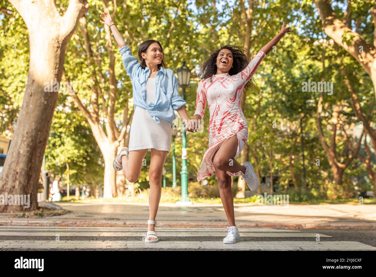 Zwei junge Frauen hüpfen freudig und halten Hände in einem lebhaften Park voller üppiger Bäume. Ihr fröhlicher Ausdruck und ihre verspielte Kleidung spiegeln einen Geist wider Stockfoto