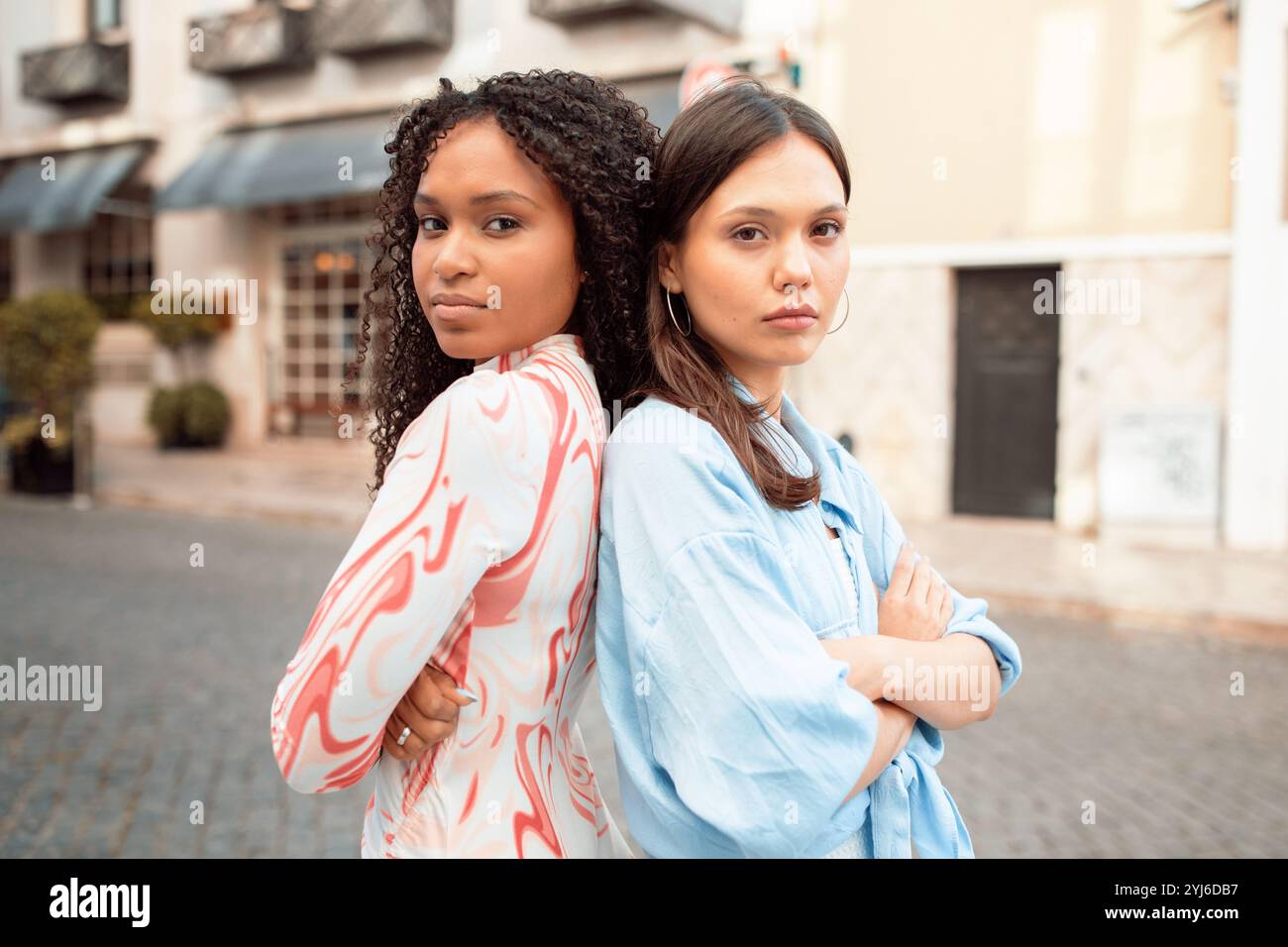 Zwei junge Frauen posieren selbstbewusst auf einer charmanten Straße. Einer hat lockige Haare und ein buntes Outfit, der andere trägt ein hellblaues Oberteil, b Stockfoto