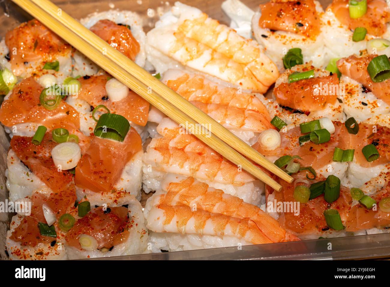 Eine Nahaufnahme eines mit Sushi gefüllten Plastikbehälters mit Lachs, Garnelen und weißem Reis mit grünen Zwiebeln und roten Chiliflocken. Holz Stockfoto