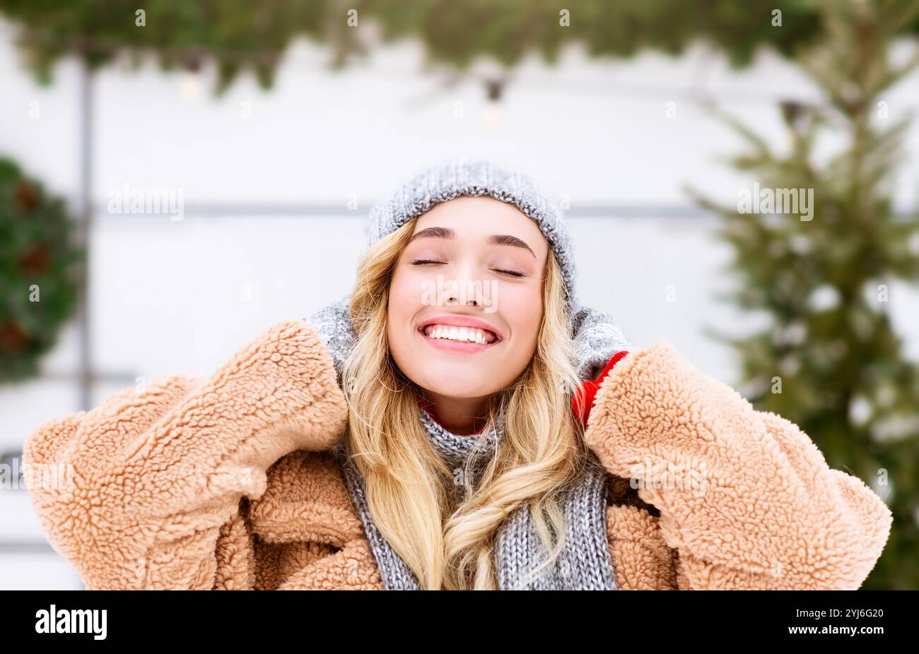 Eine junge Frau genießt einen schönen Wintertag. Sie trägt einen kuscheligen Pelzmantel und einen Strickmütze, lächelt mit geschlossenen Augen, umgeben von Schnee und Stockfoto